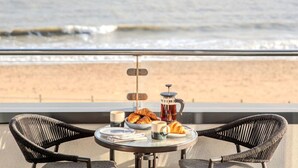 Ein Balkon mit Korbstühlen, einem Tisch mit Croissants, einer Kaffeekanne und Blick auf den Strand.