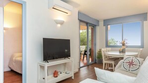Een moderne woonkamer met een flatscreen televisie op een witte standaard, een bank en een zithoek met een tafel en stoelen.