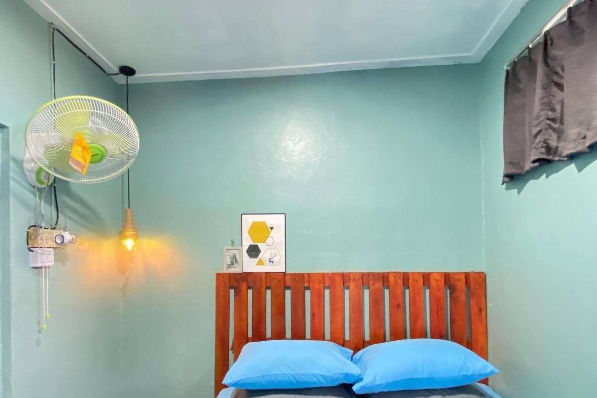 A small, neatly arranged bedroom with a single bed, a wooden headboard, a wall-mounted fan, a small table with a lamp, and a framed picture.