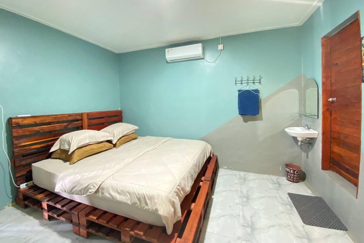 A bedroom with a wooden bed, a small bedside table, a wall-mounted air conditioner, a bathroom with a sink and mirror, and a door with a wooden frame.