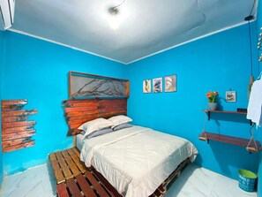 A bedroom with a blue wall, a wooden headboard, a white bed, and a small shelf with a plant. - Second Hope Lodge (Padang)