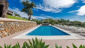 Piscina retangular com parede de pedra de um lado e pérgula com toldo branco do outro. Há espreguiçadeiras ao longo da borda da piscina.
