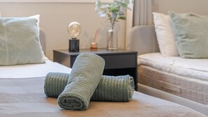 Uma cama bem arrumada com toalhas dobradas, uma mesa de cabeceira com abajur, e vista para uma janela com vegetação lá fora.
