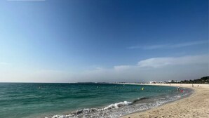 Ein Strand mit klarem, blauem Wasser, einem Sandufer und einer entfernten Küste mit Gebäuden.