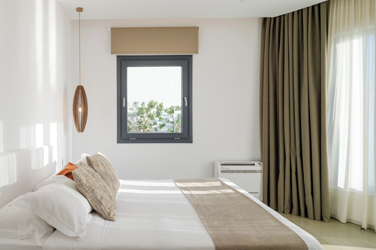 A modern hotel room with a bed, a window, a ceiling light, and a view of greenery outside.