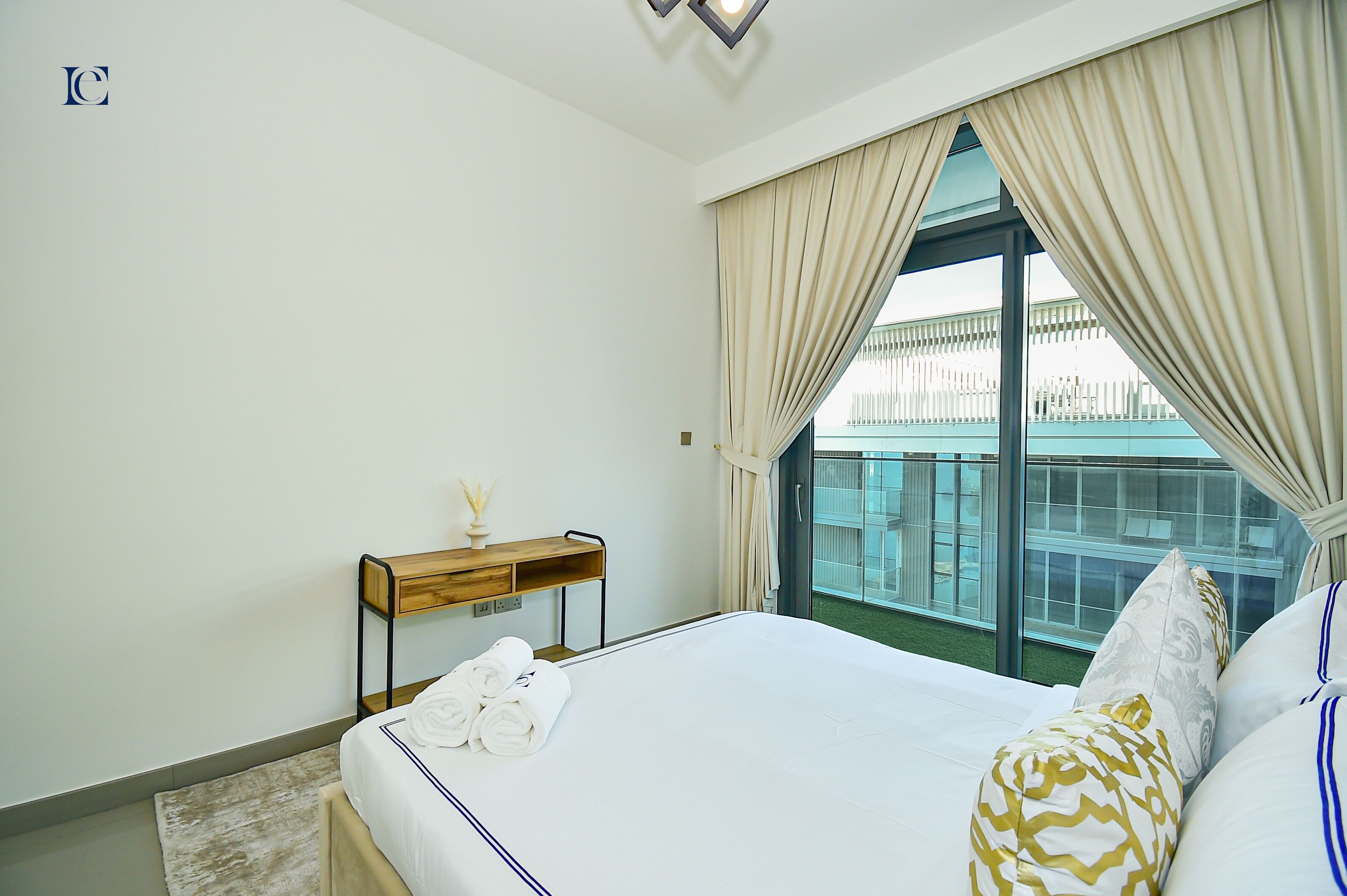 A hotel room with a large bed, a wooden bedside table with a vase, and a view of a building through the window.