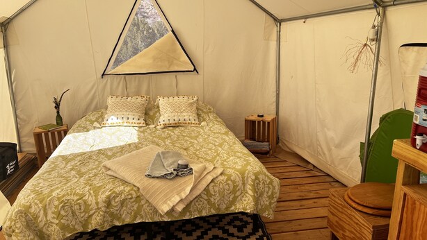 Una habitación con carpa que incluye una cama, almohadas, una colcha, un taburete de madera y una silla verde.