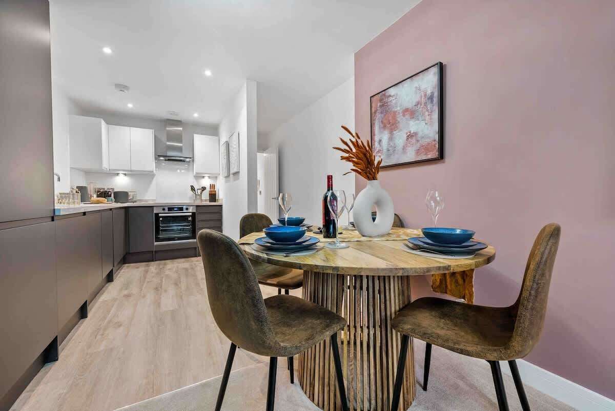 Eine moderne Küche mit weißen Schränken, ein Essbereich mit Holztisch und -stühlen sowie eine rosa Wand mit einem gerahmten Bild.