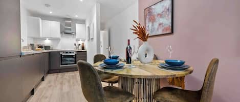 A modern kitchen with white cabinets, a dining area with a wooden table and chairs, and a pink wall with a framed picture.