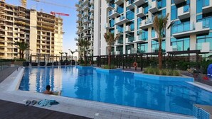 A swimming pool with a person in the water, surrounded by modern buildings and palm trees. - Burj Khalifa View Studio in Jeddaf - FAR (Dubai)