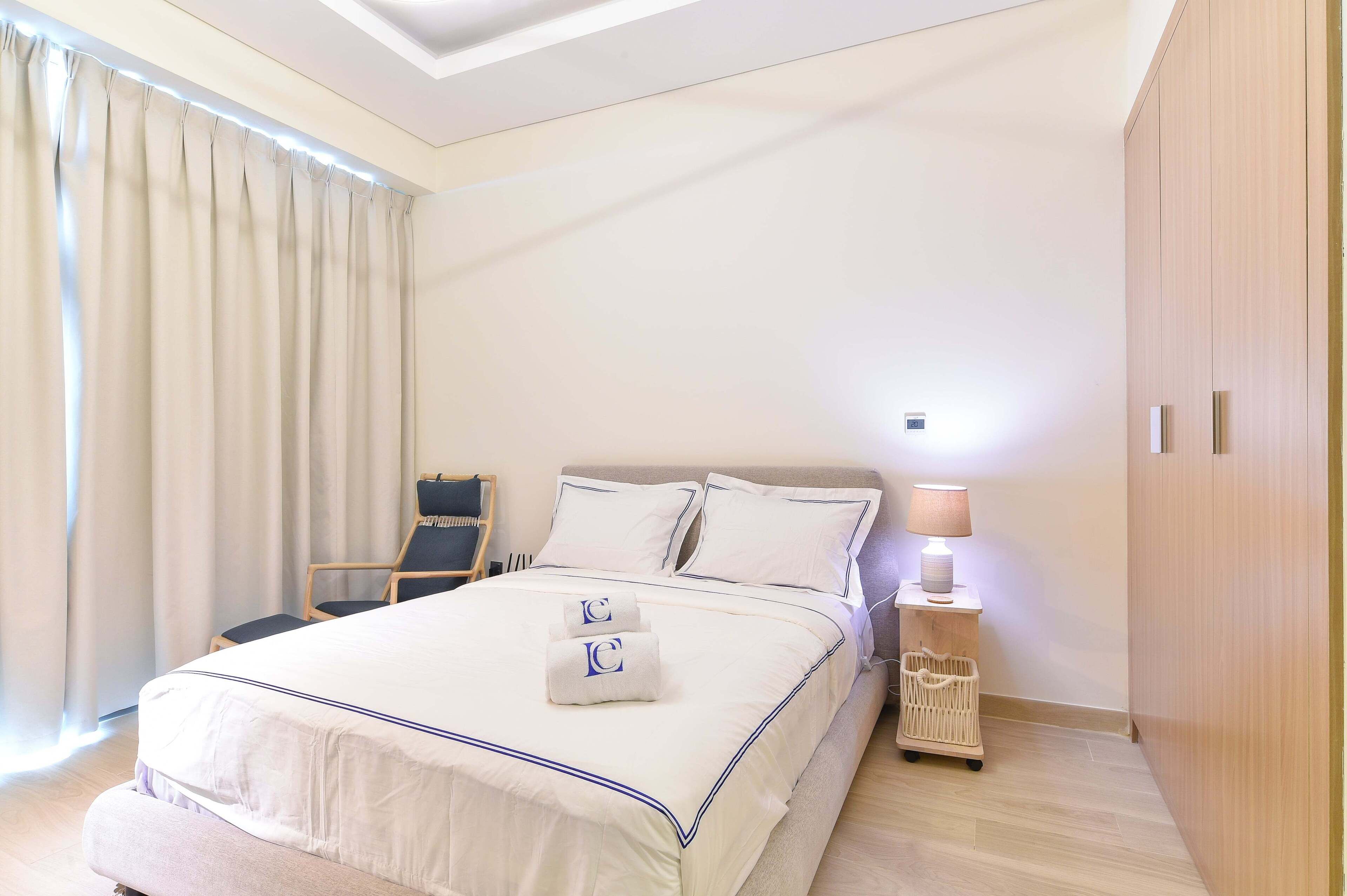 Uma cama bem arrumada com colcha e almofadas brancas, uma luminária de cabeceira, uma cadeira e um guarda-roupa de madeira em um quarto de hotel.