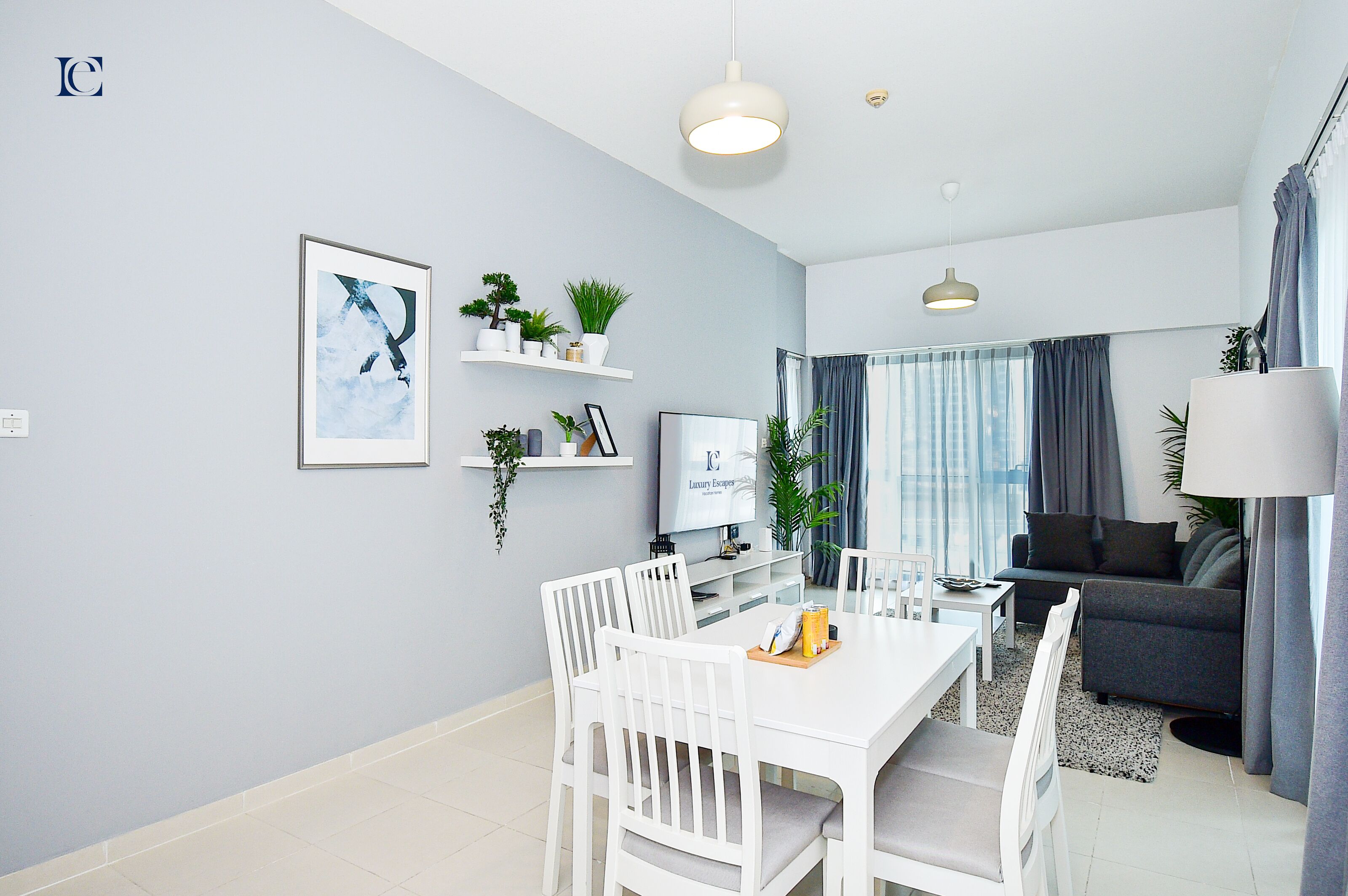 Ein modernes Wohnzimmer mit grauem Sofa, einem Esstisch mit Stühlen und einem Fernseher auf einem Schrank. Pflanzen stehen auf Regalen und ein großes Fenster mit Vorhängen ist zu sehen.