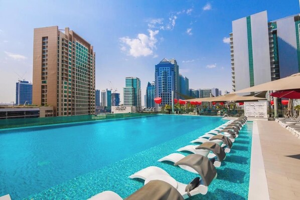 A hotel pool with lounge chairs, a city skyline in the background, and a clear blue sky.
