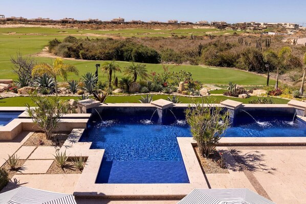 Una piscina con caída de agua, rodeada de jardines y palmeras.