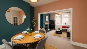 A dining area with a round table set for four, surrounded by chairs. There is a mirror on the wall, a fireplace, and a sofa in the background.