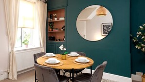 A dining area with a round table set for four, surrounded by dark chairs. A mirror hangs on the teal wall, reflecting part of the room.