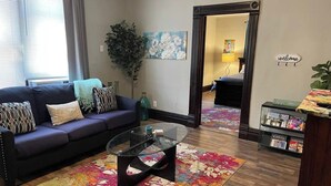 Una sala de estar con sofá, mesa de centro y una alfombra colorida. Hay una planta grande, un cuadro y un cartel con la palabra 'welcome'.