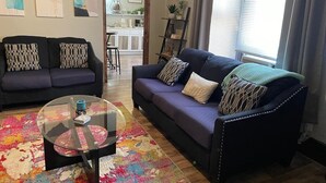 Un salón con dos sofás oscuros, una mesa de centro de vidrio y una alfombra colorida.
