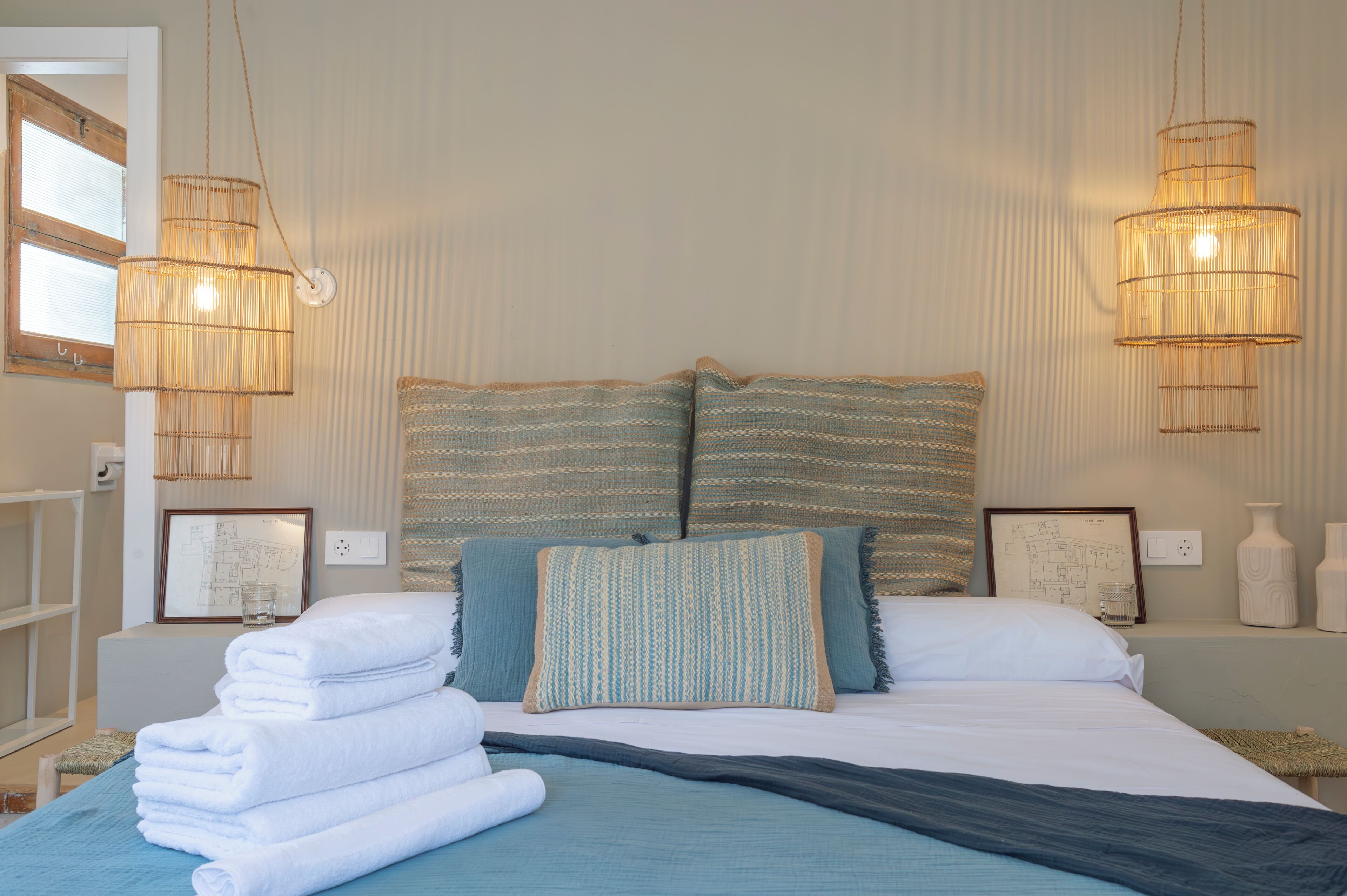 A neatly made bed with folded white towels, two large striped pillows, and a blue blanket. There are two wall-mounted lamps with a woven design.