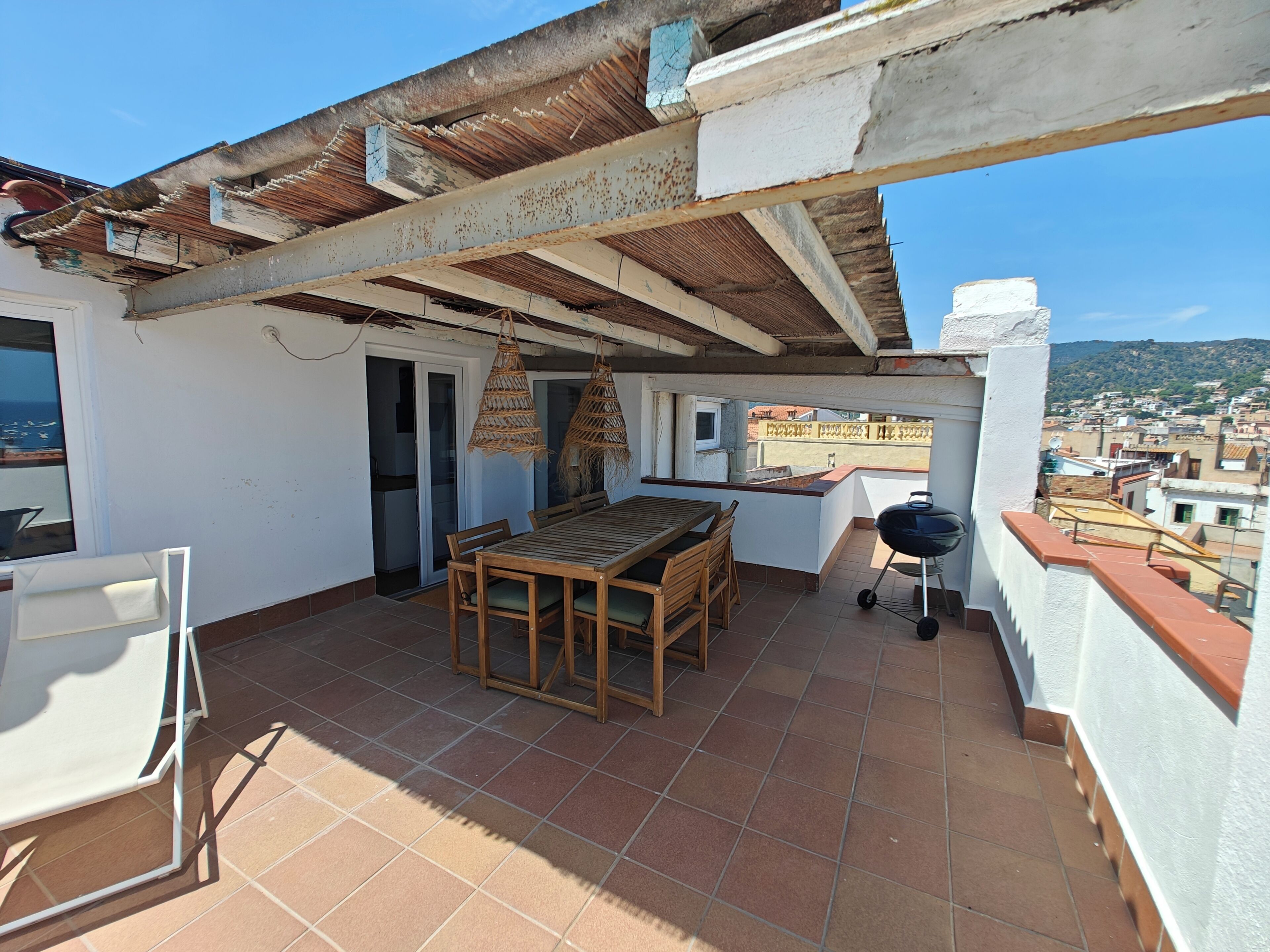 A rooftop terrace with a wooden table and chairs, a grill, and a view of the city.