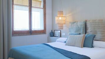 Un dormitorio con una cama grande, una lámpara colgante, una ventana con persianas y una mesita de noche con un jarrón y un cuadro enmarcado.