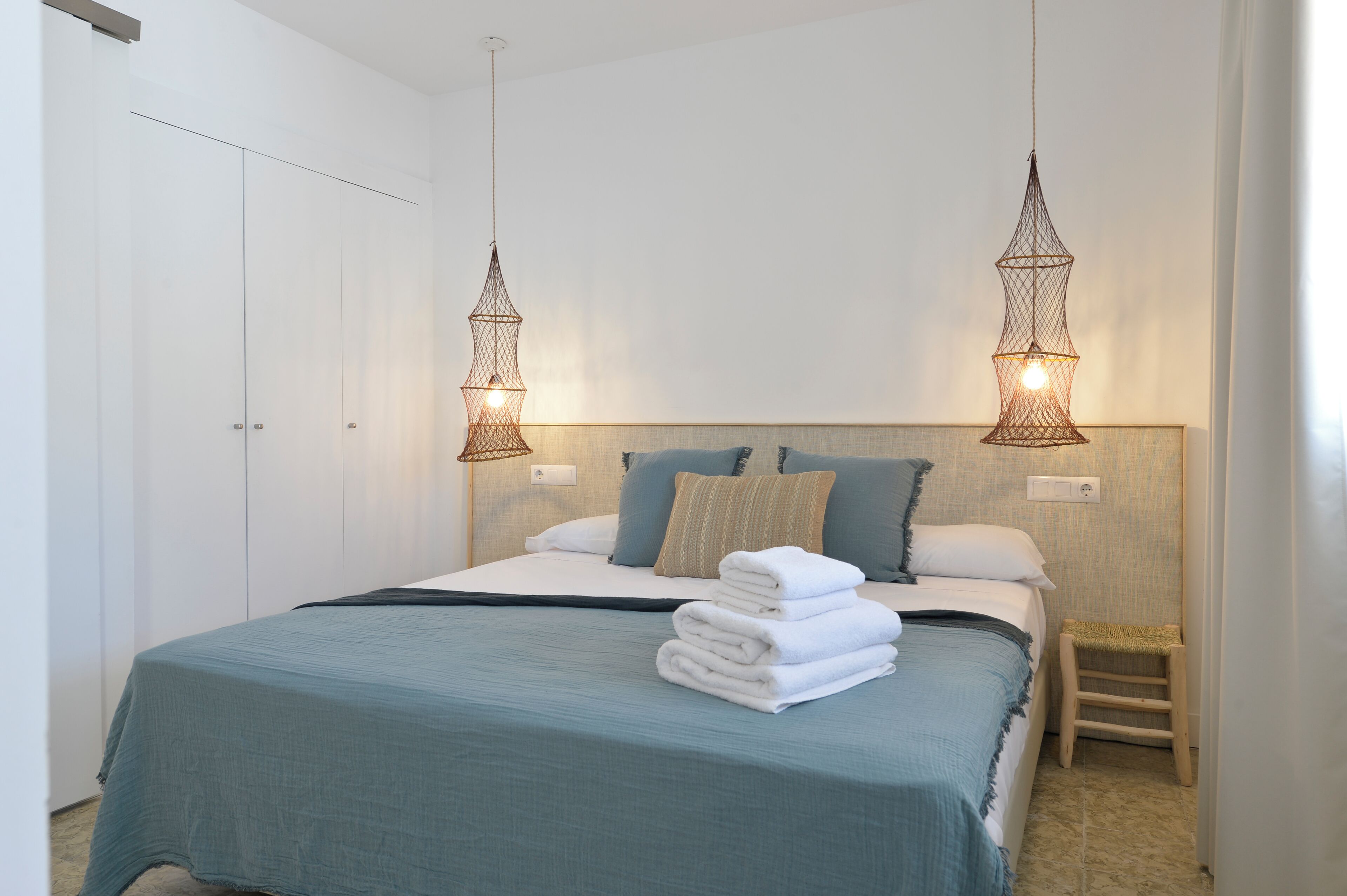 A neatly made bed with a blue bedspread, white pillows, and a stack of white towels. There are two hanging light fixtures above the bed.