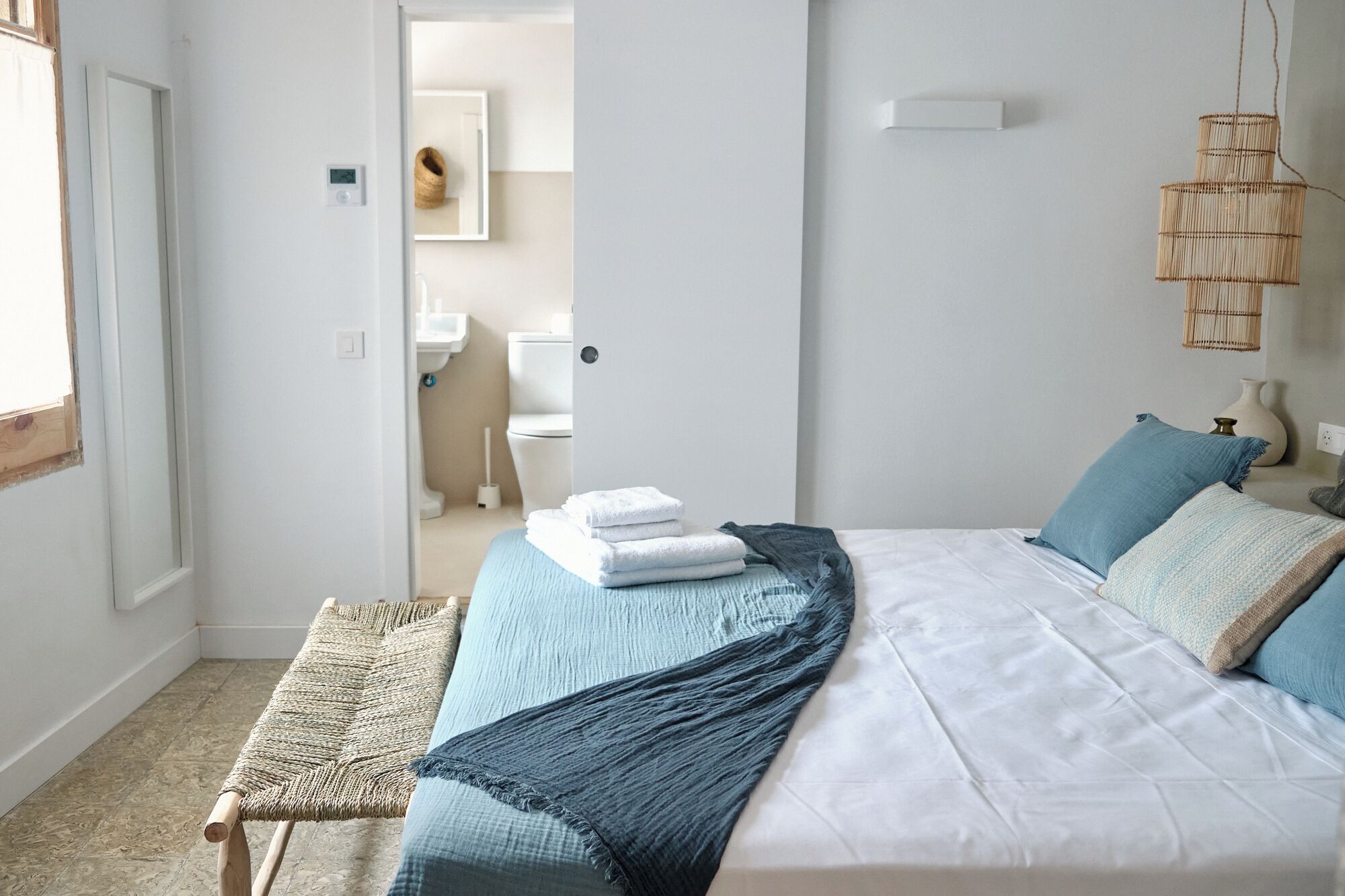 A bedroom with a bed, a bedside table, a woven bench, and a view into the bathroom.