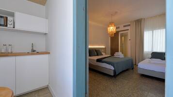 A hotel room with a kitchenette, a bed with a blue bedspread, a bedside table, and a large window with curtains.