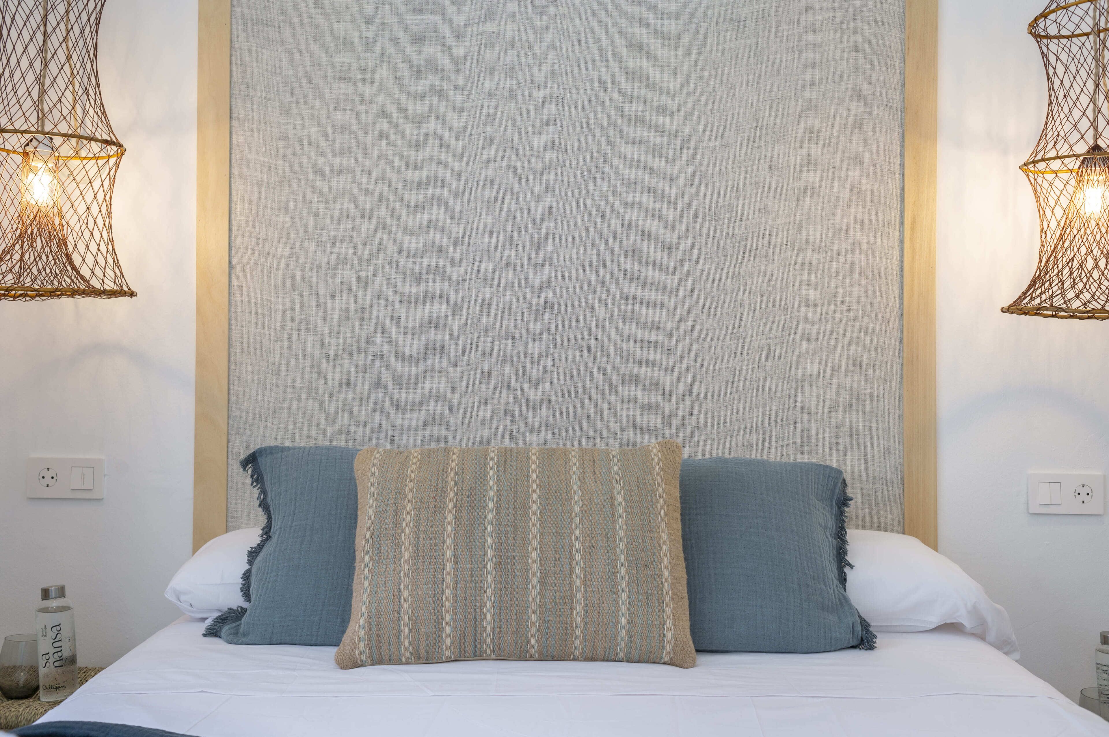 Una cama bien hecha con dos almohadas, una pared con textura y dos lámparas fijadas a la pared.