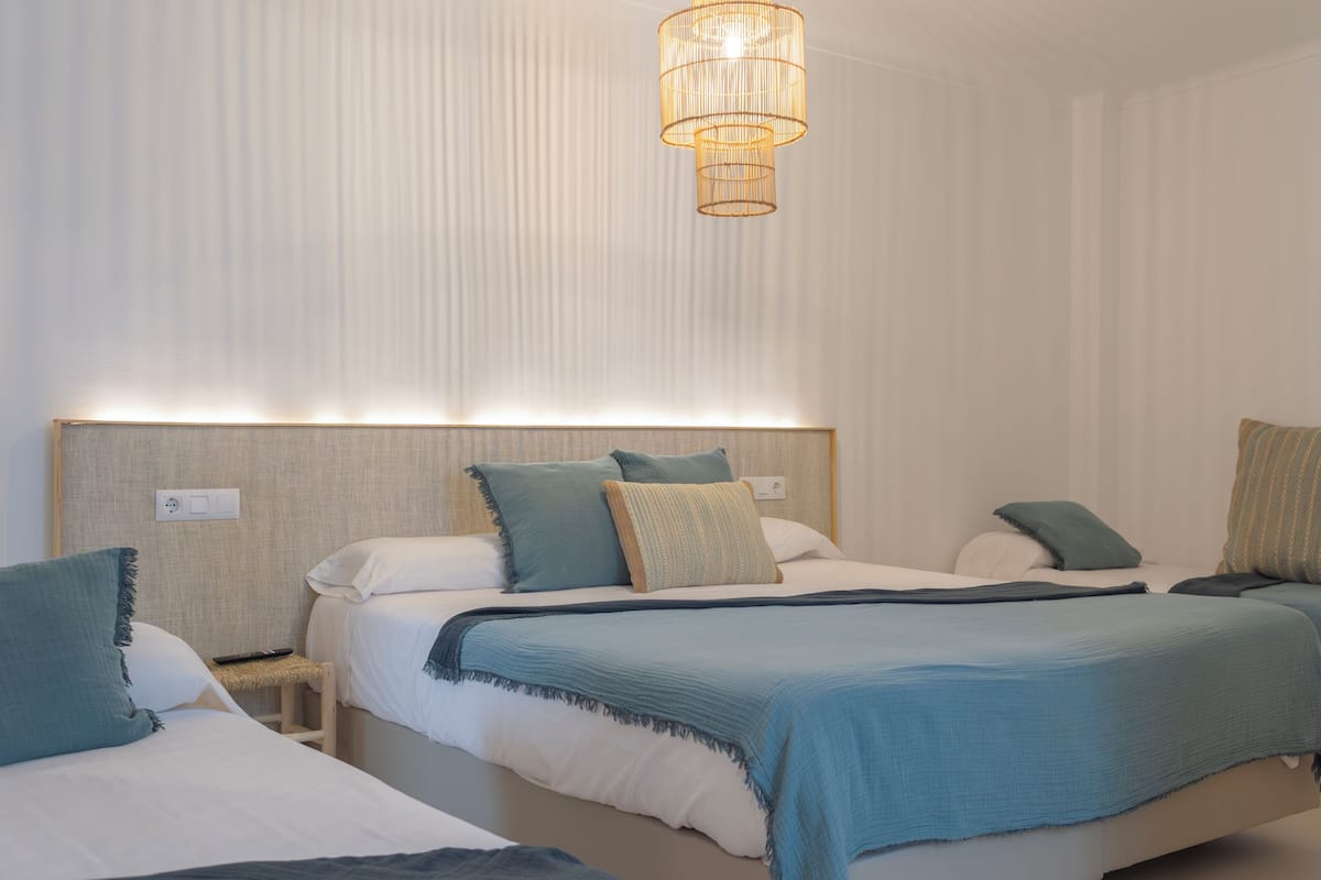 Hotellihuone, jossa on kolme sänkyä, jokaisessa sängyssä on pääty ja huoneessa on riippuvalaisin.