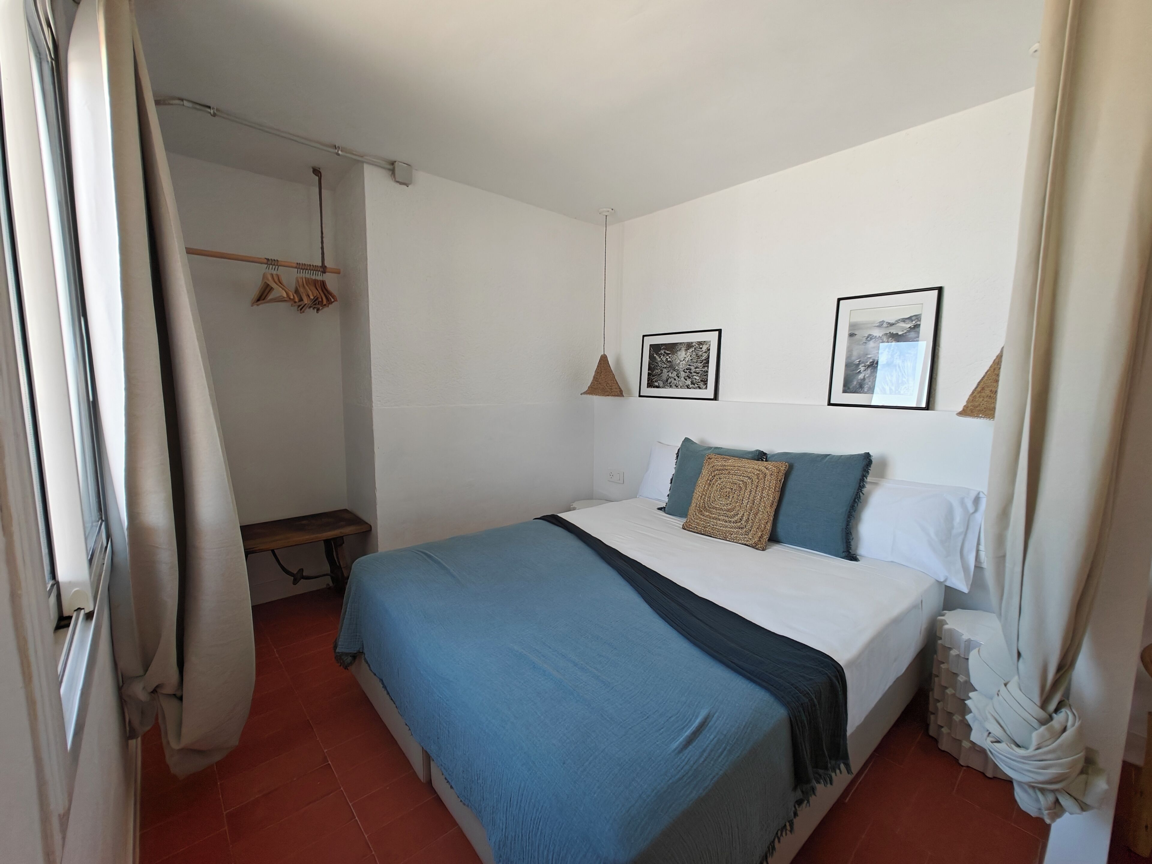 Quarto com cama, luminária pendente, um banquinho e um cabideiro fixado na parede.