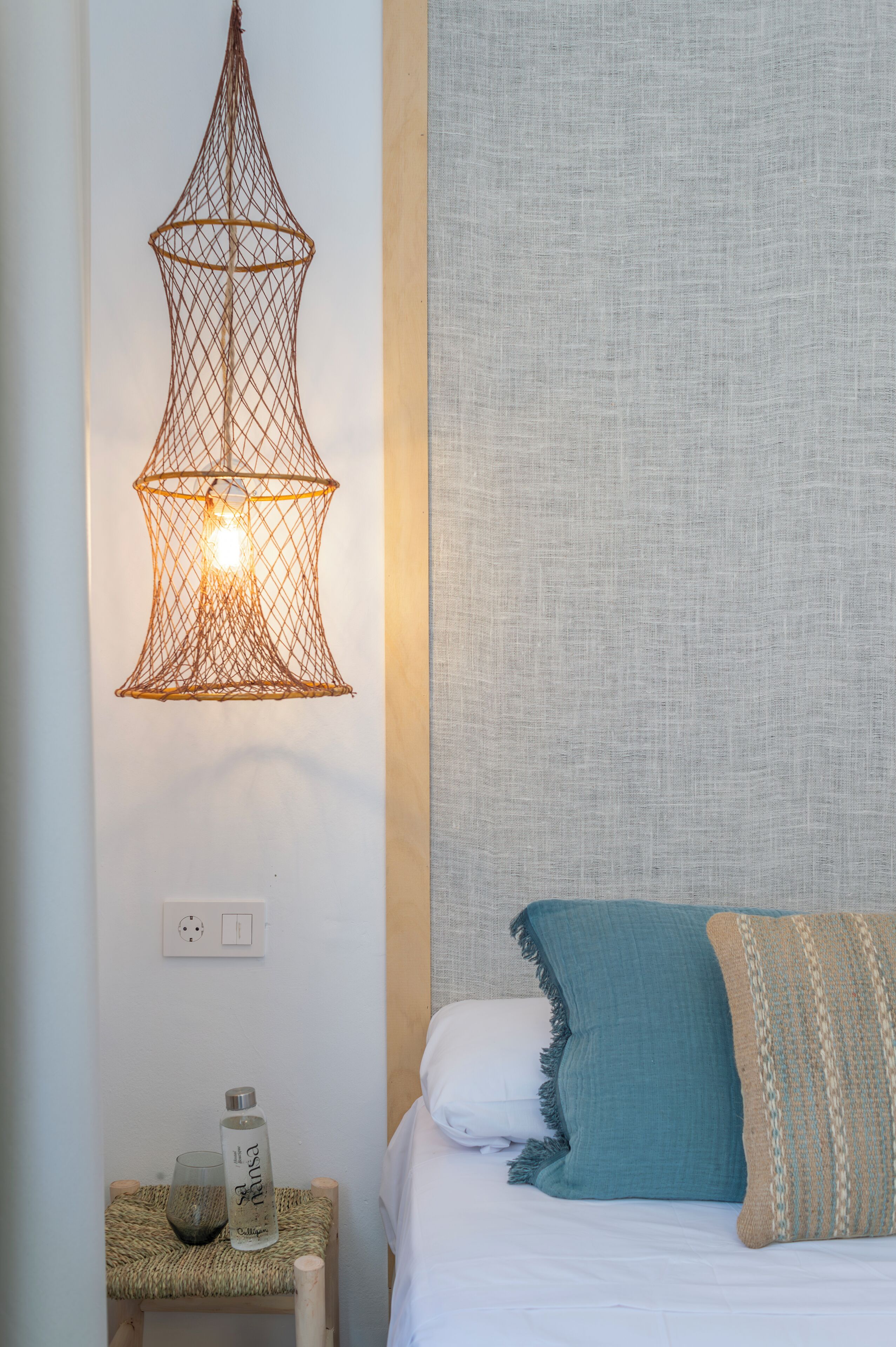 Een netjes opgemaakt slaapkamer met een bed, een wandlamp en een tafeltje met een fles en een glas.