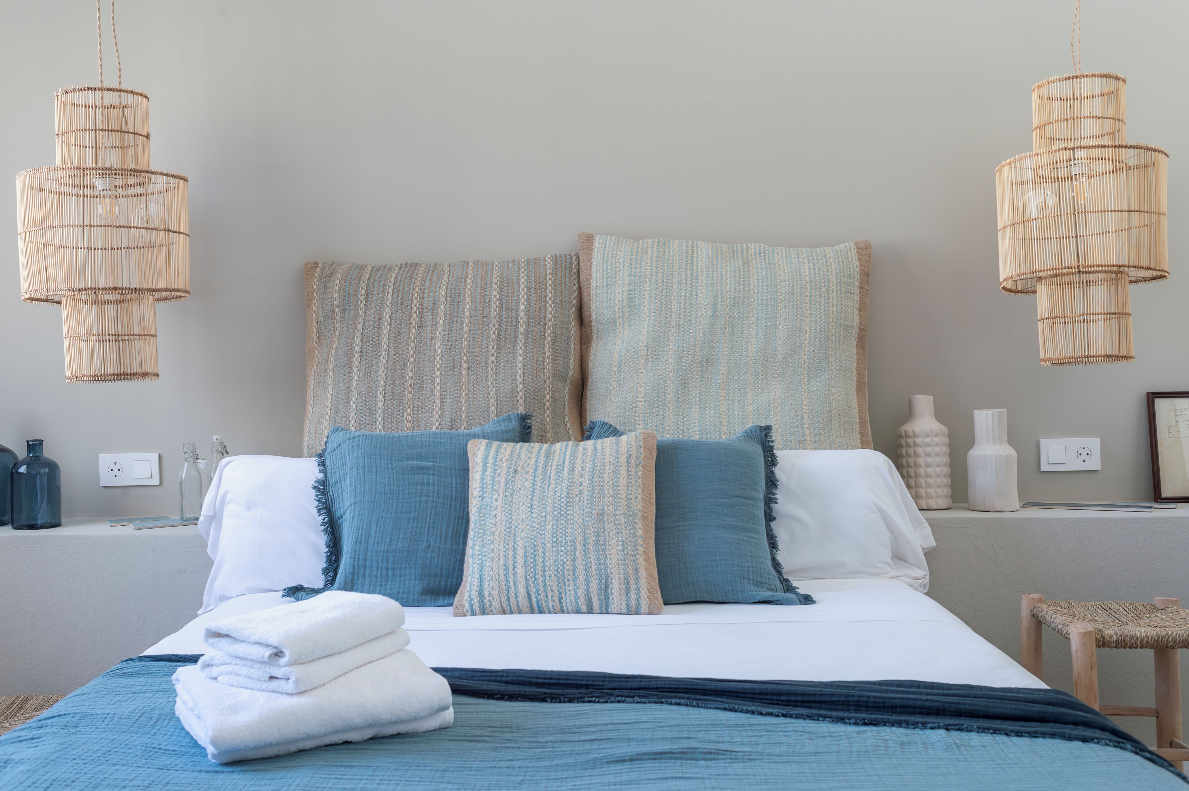 Una cama bien hecha con ropa de cama azul y blanca, dos lámparas de mimbre colgantes y un taburete pequeño de madera.