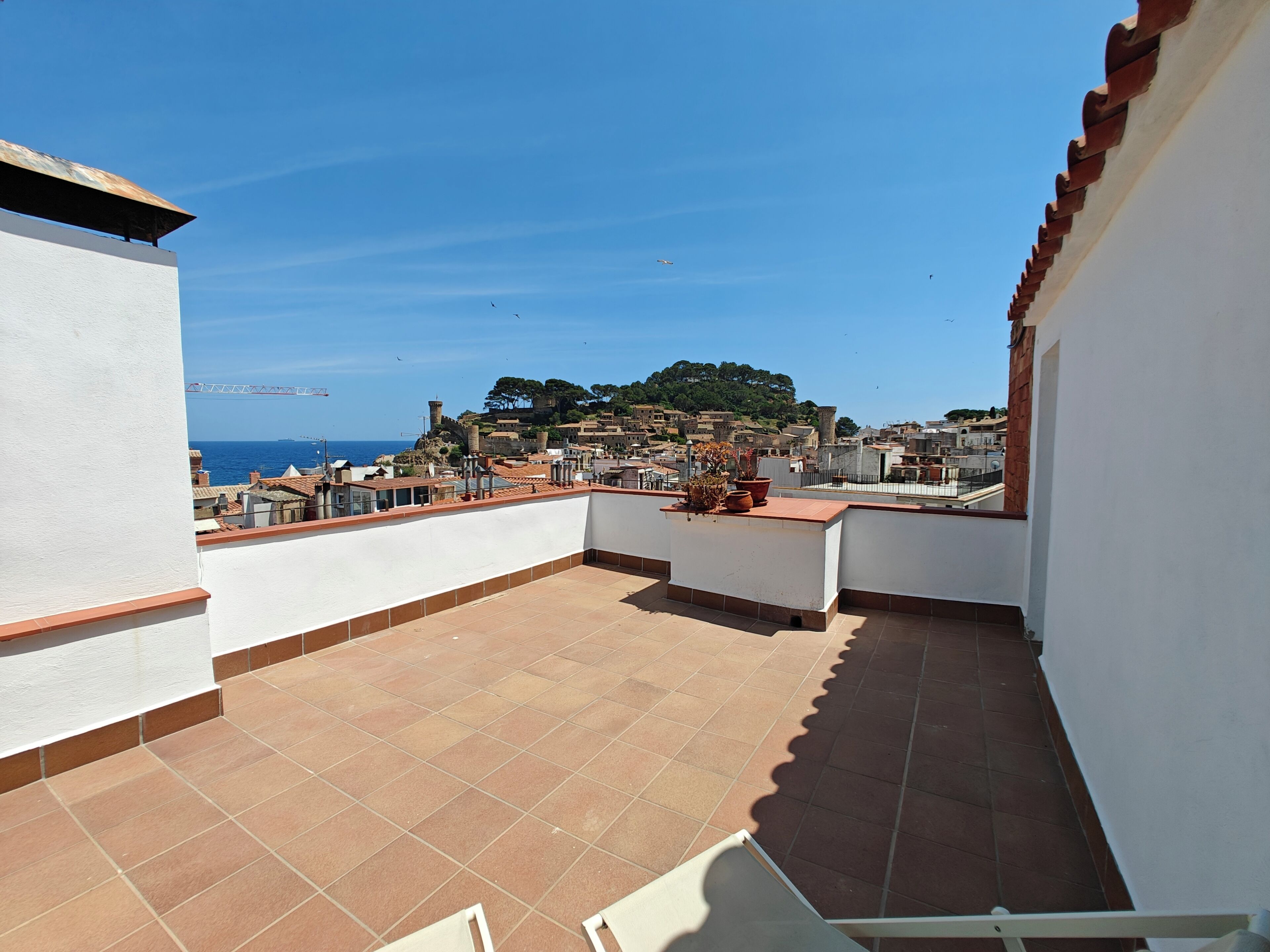 A rooftop terrace with a view of a coastal town, terracotta tiles, and a white wall.