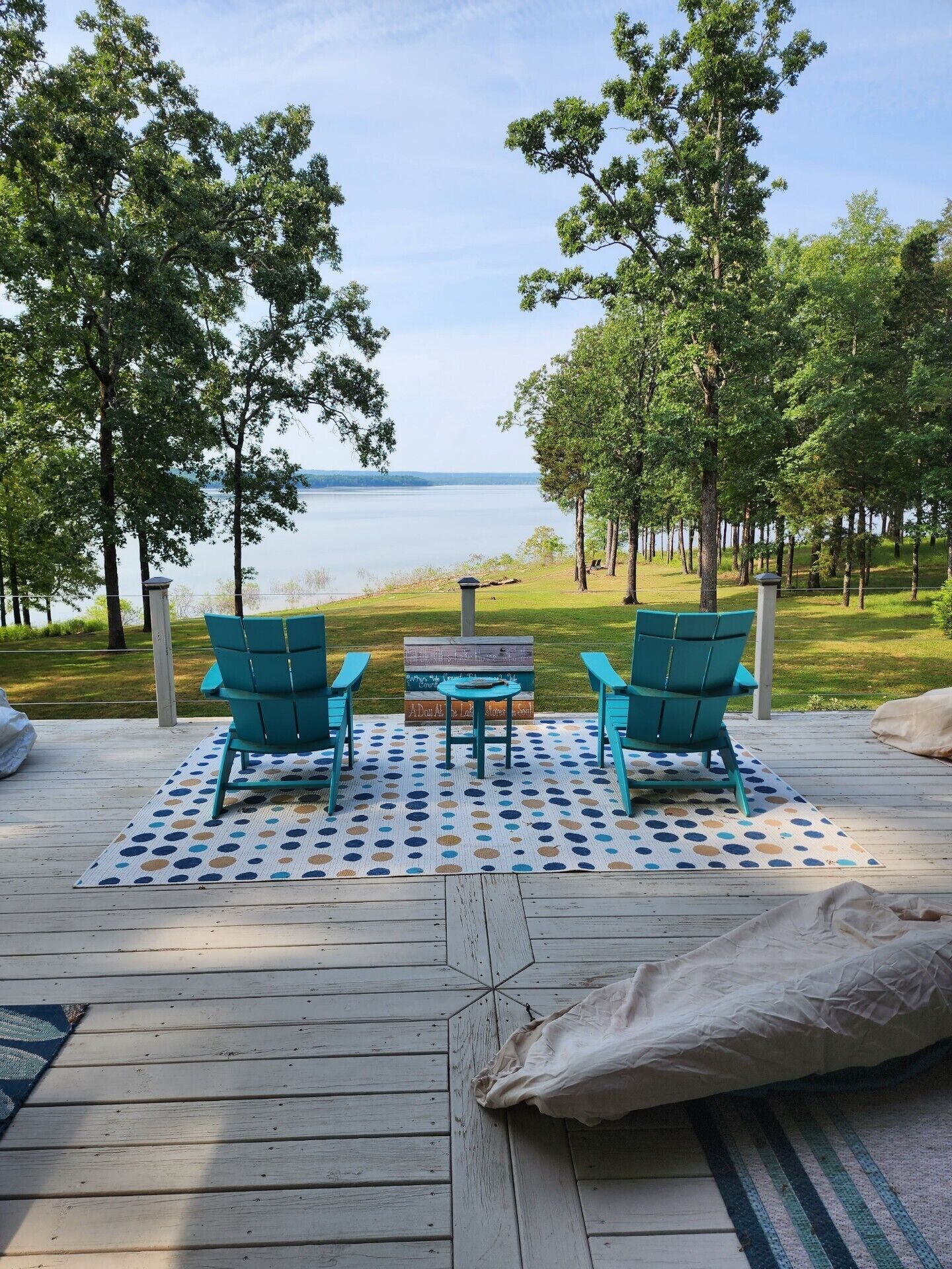A deck with two turquoise Adirondack chairs, a small table, and a view of a lake.
