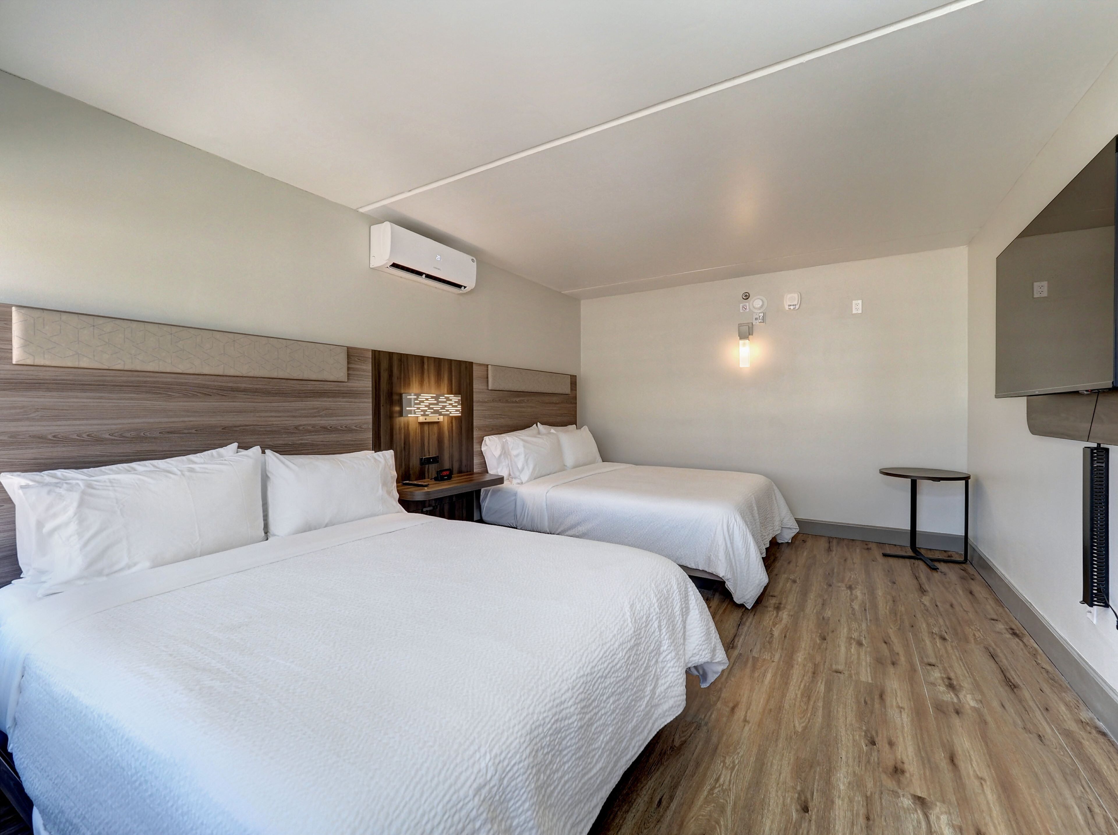 A hotel room with two beds, a wooden headboard, a wall-mounted air conditioner, a small table, and a wall-mounted TV.