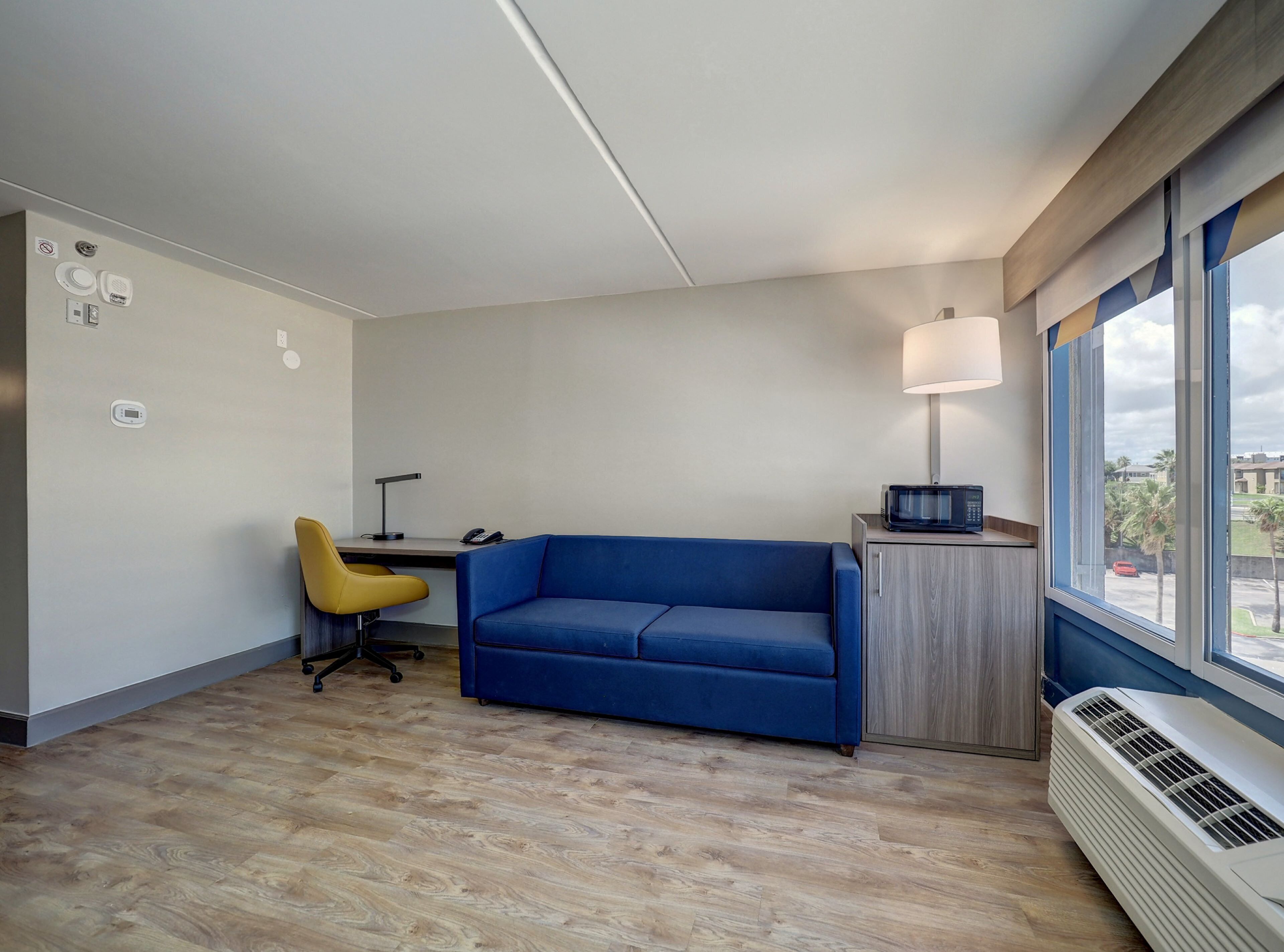 Une pièce moderne avec un canapé bleu, un fauteuil jaune, un bureau avec une lampe et une fenêtre donnant sur des palmiers.