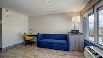 Une pièce moderne avec un canapé bleu, un fauteuil jaune, un bureau avec une lampe et une fenêtre donnant sur des palmiers.