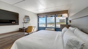 A hotel room with a bed, a desk, a chair, a TV, and a large window with a view of buildings and a clear sky. - Holiday Inn Express Corpus Christi – Beachfront by IHG (Corpus Christi)