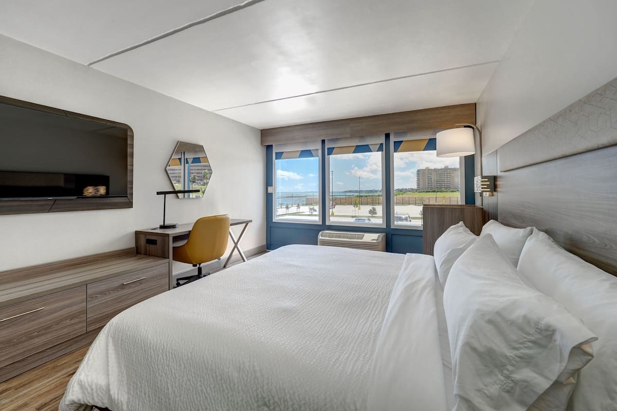 A hotel room with a bed, a desk, a chair, a TV, and a large window with a view of buildings and a clear sky.