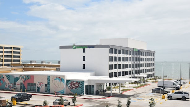 A modern hotel with a white facade, a Holiday Inn Express sign, and a mural on the side. - Holiday Inn Express Corpus Christi – Beachfront by IHG (Corpus Christi)