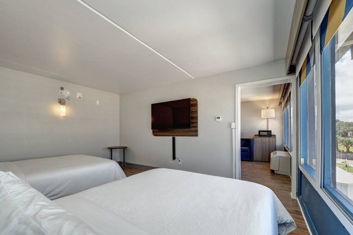 A hotel room with two beds, a TV mounted on the wall, a small table, and a view of the outdoors through a window.