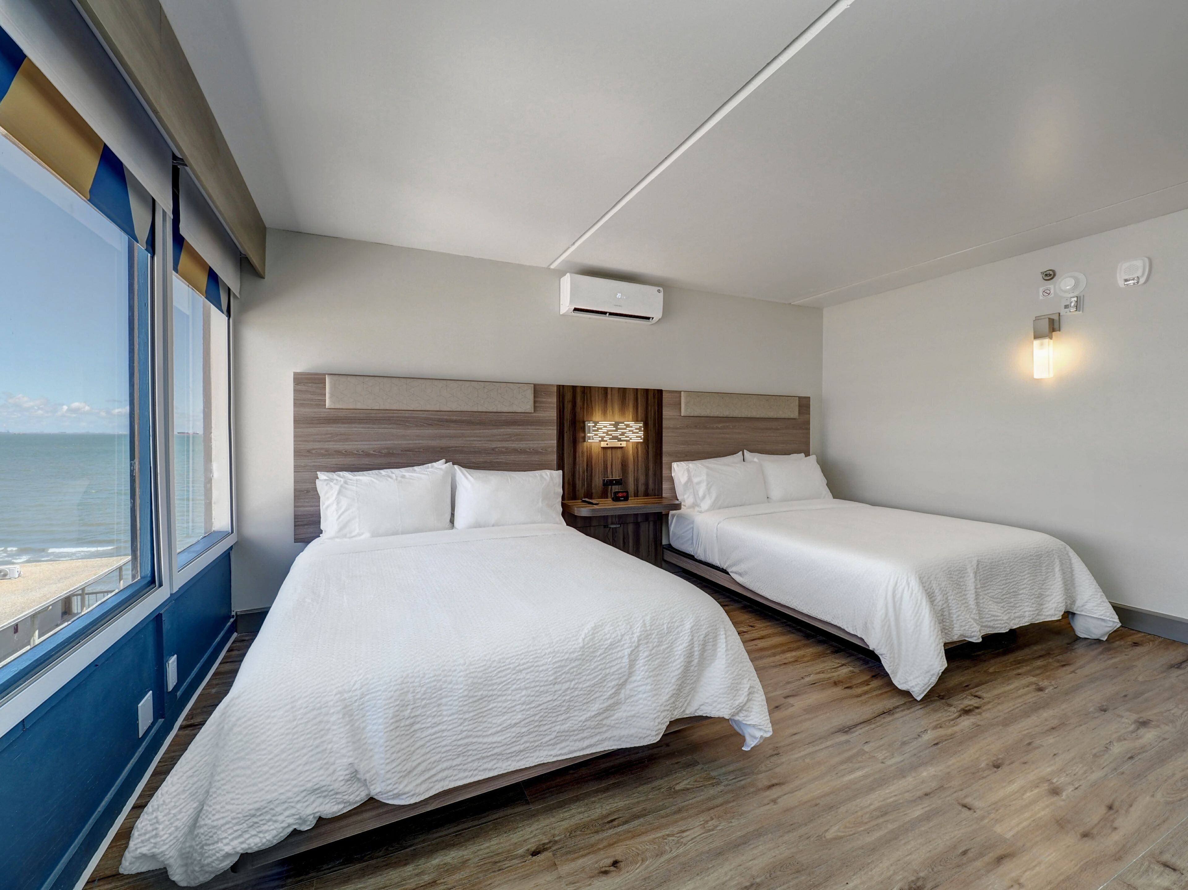 A modern hotel room with two beds, a wooden headboard, a nightstand, and a view of the sea.