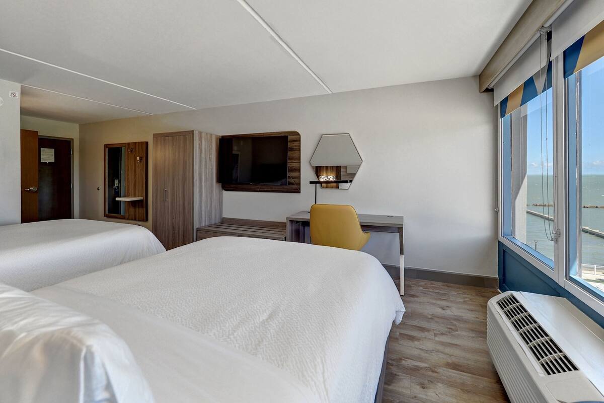Quarto de hotel com duas camas, uma escrivaninha com cadeira, uma televisão e uma janela grande com vista.