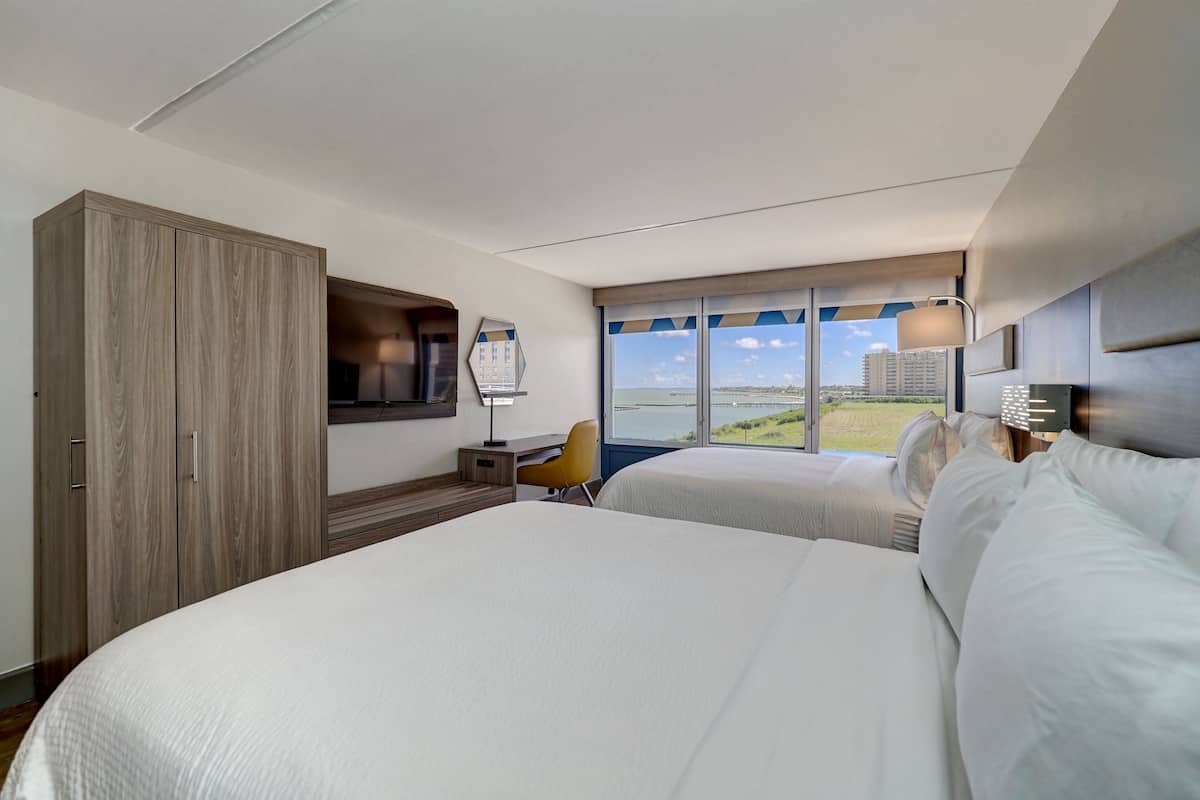 A hotel room with two beds, a desk, a chair, a large wardrobe, and a view of the sea.