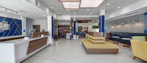 A modern hotel lobby with a reception desk, seating area, and a visible store in the background.