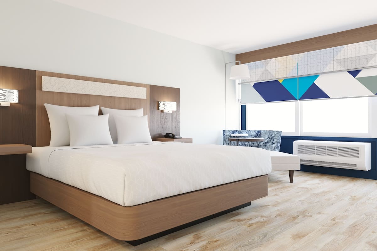 A modern bedroom with a wooden headboard, a large bed with white bedding, bedside lamps, a nightstand, a small table with a chair, and a window with a geometric view.