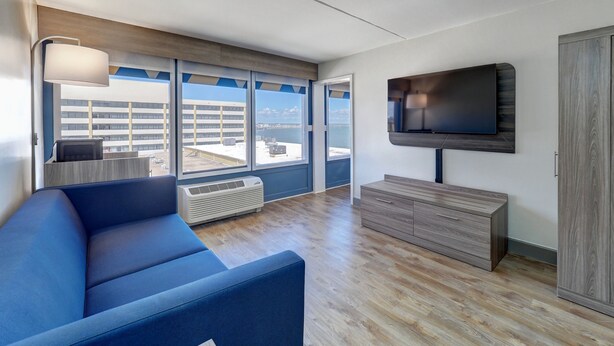 Un salon moderne avec un canapé bleu, un téléviseur à écran plat fixé au mur et de grandes fenêtres donnant sur le paysage urbain.