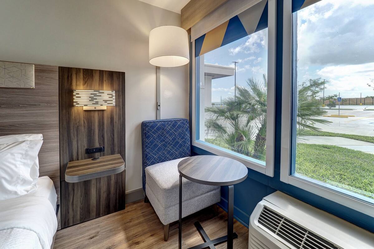 Quarto de hotel moderno com cama, uma mesinha, uma cadeira, janela com vista para o exterior e luminária na parede.