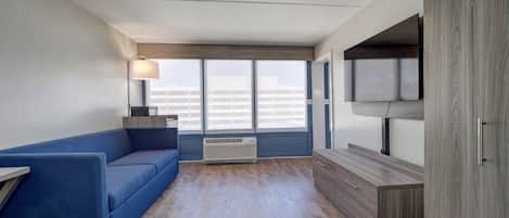 A modern hotel room with a blue sofa, a desk with a lamp, a TV mounted on the wall, and a large window with a view of buildings.